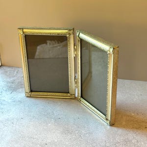 May include: A hinged double picture frame featuring two rectangular openings with ornate gold-toned metal borders. The frames are connected by a central metal hinge, allowing the unit to stand upright in a folded, V-shaped configuration.