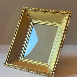 May include: A gold-colored rectangular picture frame with a decorative rope-like border and corner leaf accents. The frame has a glass front and a cardboard backing, suitable for displaying photographs or artwork. The frame is angled slightly.