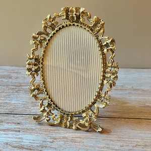 May include: Ornate gold-colored oval picture frame with floral and scroll detailing. The frame has a stand and a cream-colored, vertically striped backing. A decorative accent for displaying photos or artwork.