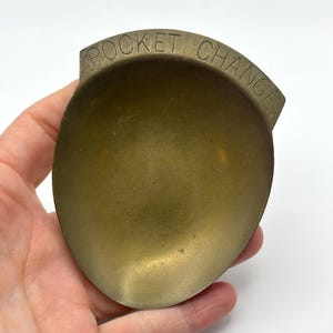 May include: A vintage brass pocket change dish. The dish is oval-shaped with a curved top, and the words "POCKET CHANGE" are embossed across the top. The dish has a smooth, polished surface and a warm, golden color.