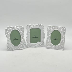 May include: Three white Hallmark picture frames with ornate floral and scroll designs. One frame is oval, and two are rectangular. Each frame has a green interior and the Hallmark logo.