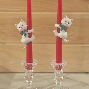 May include: Two red taper candles with white cat figurines and clear crystal candle holders. The cats have green and red accents. The candle holders have a detailed floral design.