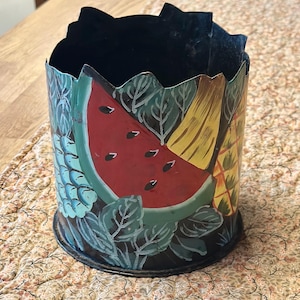 May include: A cylindrical metal container with a black interior and a colorful exterior featuring painted fruits. The design includes a watermelon slice, pineapple, and grapes, with green leaves. The container sits on a patterned surface.