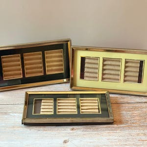 May include: Three vintage jewelry display cases with gold frames. Each case has three rectangular compartments with a beige, ribbed interior. The cases have black, yellow, and green backgrounds.