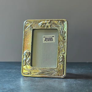 May include: A rectangular silver-plated picture frame featuring embossed floral and dragonfly motifs. The frame has a textured, hammered finish and includes a central sticker with the text GENUINE SILVER PLATED in black capital letters.
