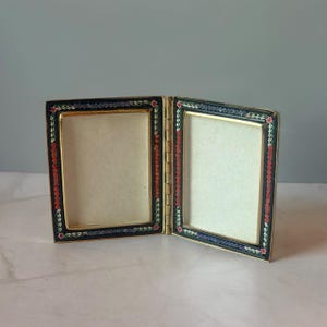 May include: A hinged double photo frame with a gold-tone metal finish. The rectangular borders feature a black enamel inlay decorated with a repeating pattern of small red flowers and green leaves, creating a colorful floral motif.