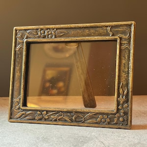 May include: A rectangular picture frame with a metallic bronze finish and a weathered, antique appearance. The frame features raised relief detailing of floral motifs and flowing vine patterns along all four edges, surrounding a clear glass pane.