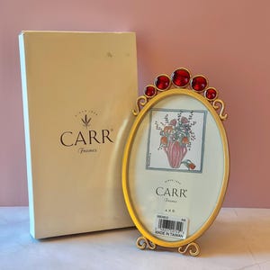 May include: A gold-colored oval picture frame with red jewel accents and a decorative base. The frame holds a floral print and is accompanied by a cream-colored box. The frame is labeled "CARR Frames".
