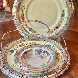 May include: A silver-plated serving tray with a handle, featuring a floral design on a yellow background. The tray has a decorative rim and holds a smaller dish with a mirrored center. A matching plate is in the background.