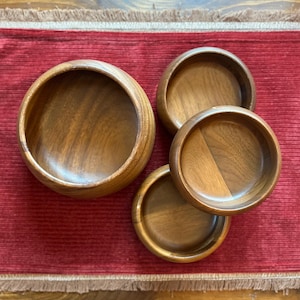 May include: Four wooden bowls of varying sizes are arranged on a red surface. The bowls are made of a light brown wood with visible grain patterns. The largest bowl is on the left, with three smaller bowls to the right.