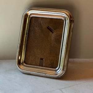 May include: A rectangular silver-coloured picture frame with rounded corners. The frame has a shiny, metallic finish and a dark brown backing. The frame is designed to hold a photograph or artwork and is suitable for home decor.