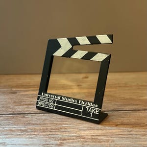 May include: A black and white film clapperboard picture frame featuring the text Universal Studios Florida, PROD NO, DIRECTOR, and TAKE 1. The frame has a classic diagonal striped top bar and a rectangular opening for a photograph.