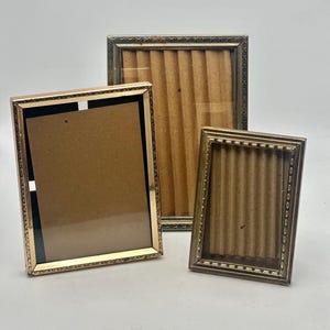 May include: Three vintage picture frames in different sizes. The frames have a gold-coloured metal finish with decorative detailing around the edges. The frames are empty, with a cardboard backing visible.