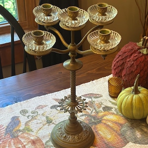 May include: A five-arm brass candelabra featuring a central twisted stem, a decorative circular base with ornate detailing, and five clear glass bobeches with fluted edges. Each arm holds a brass candle cup designed to support a single taper candle.