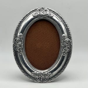 May include: Oval silver-toned picture frame with ornate floral and scroll detailing. The frame has a brown backing and is designed to hold a photograph. A decorative piece for displaying cherished memories.
