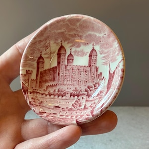 May include: A small, round ceramic dish featuring a detailed red and white transferware print of the Tower of London. The design depicts the historic castle architecture with turrets and battlements against a cloudy sky, rendered in a monochromatic red ink.