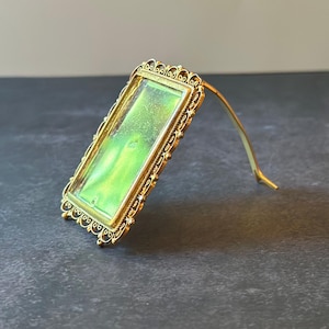 May include: A rectangular gold-toned frame featuring an intricate filigree border and a vibrant, shimmering lime green insert. The frame is supported by a thin, curved gold-toned metal stand attached to the back, allowing it to stand upright.