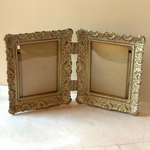 May include: A gold-colored, hinged double picture frame with ornate, scrolling details. The frame is open, revealing two empty spaces for photos. The frame is made of a metallic material and has a vintage aesthetic.