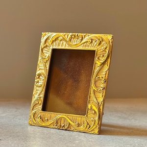 May include: A rectangular picture frame with a metallic gold finish, featuring an ornate, raised relief pattern of scrolling acanthus leaves and sunburst-like rays surrounding a dark, solid brown center panel within the frame's inner border.