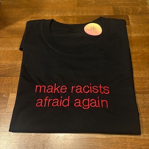 Könnte beinhalten: Schwarzes T-Shirt mit dem gestickten Schriftzug "make racists afraid again" in Rot. Das Shirt ist sauber auf einer Holzoberfläche gefaltet. Ein kleiner Aufkleber ist in der Nähe des Halsausschnitts sichtbar.