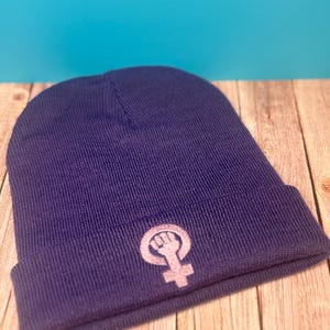 Könnte beinhalten: Eine dunkelviolette Mütze mit einem hellrosa gestickten feministischen Symbol. Die Mütze ist aus einem Strickmaterial gefertigt und wird auf einer Holzoberfläche mit blauem Hintergrund präsentiert. Das Symbol zeigt eine geballte Faust innerhalb eines Frauensymbols.