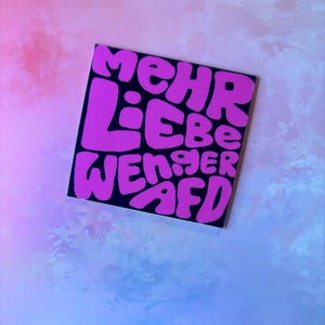 Könnte beinhalten: Ein quadratischer Aufkleber mit den Worten "mehr Liebe weniger AfD" in Buchstaben. Der Text ist leuchtend rosa auf schwarzem Hintergrund. Der Aufkleber befindet sich auf einem Hintergrund aus Rosa- und Blautönen.