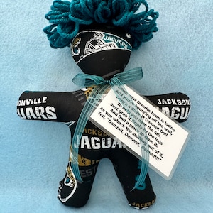 May include: A fabric doll featuring a black, teal, and gold Jacksonville Jaguars print with teal yarn hair. A teal ribbon is tied around the neck, holding a clear tag with the text: When your favorite team is losing And you're bouncing off the wall, Here's a Slam-It Dammit Doll To help you ease the fall. Just grab it firmly by the legs And find a place to slam it. As you whack the stuffing out of it, Yell, "Dammit, Dammit, Dammit!!"