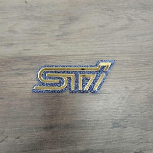 May include: A gold-colored STi emblem with a blue glitter background. The emblem is a stylized logo, likely for a car or automotive product. The letters are bold and outlined in gold, with a blue glitter fill.