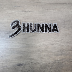 May include: A black and silver glittered "3 Hunna" decorative item. The word "Hunna" is in bold, block letters, and the number "3" is stylized with a curved design. The item is set against a light gray wood-grain background.