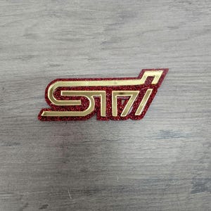 May include: A red and gold STI emblem with a glittery finish. The emblem is in a stylized font and features a gold outline. The background is a gray wooden surface.