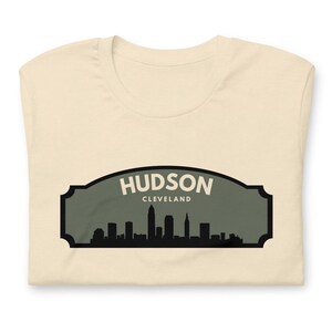 Puede incluir: Camiseta crema doblada con un gráfico del horizonte de Hudson, Cleveland. El diseño está enmarcado en un marco arqueado verde oscuro, con el nombre de la ciudad en letras blancas.