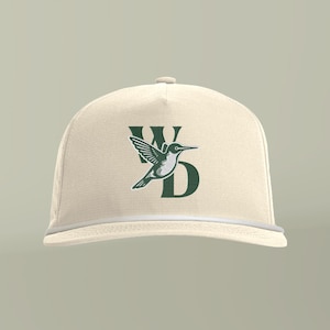 Könnte beinhalten: Eine cremefarbene Baseballkappe mit einem dunkelgrünen gestickten Logo auf der Vorderseite. Das Logo besteht aus den Buchstaben W und D in einer Serifenschrift mit einer stilisierten Kolibrigrafik, die mittig zwischen den Buchstaben platziert ist.
