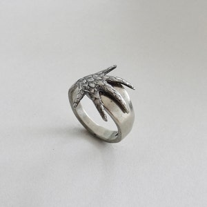 May include: A silver ring featuring a detailed, textured design of a bird's foot.