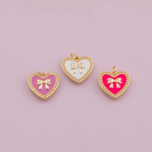 May include: Three heart-shaped pendants with gold-colored trim and bows. One pendant is pink, one is white, and one is a bright pink color. The pendants are on a light pink background.