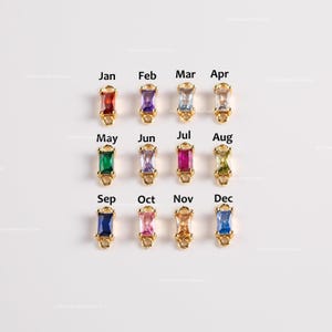 May include: A collection of twelve gold-toned charms, each featuring a rectangular gemstone in a different color. Each charm is labeled with a month of the year, from January to December, with the text in black. The gemstones are in various colors, including red, purple, blue, and green.