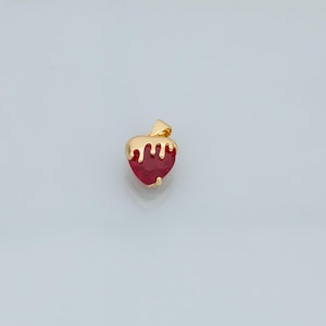 May include: A gold heart-shaped pendant with a red gemstone. The gold is dripping down the sides of the heart.