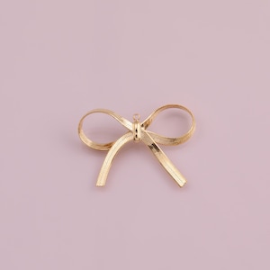 May include: A gold-colored bow pendant, crafted from a flat, woven material, lies centered against a soft pink background. The bow features a small loop at the top for hanging. The design is simple and elegant.