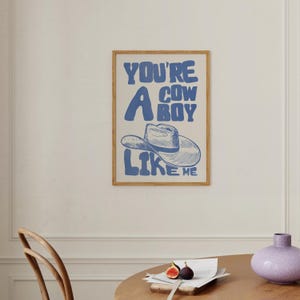 Puede incluir: Un estampado enmarcado con el texto "YOU'RE A COWBOY LIKE ME" en azul, con una ilustraci&oacute;n de un sombrero de vaquero. El estampado tiene un fondo beige claro y un marco de madera. La obra de arte se exhibe en una pared blanca.