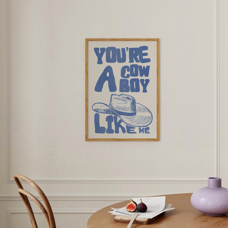 Puede incluir: Un estampado enmarcado con el texto "YOU'RE A COWBOY LIKE ME" en azul, con una ilustraci&oacute;n de un sombrero de vaquero. El estampado tiene un fondo beige claro y un marco de madera. La obra de arte se exhibe en una pared blanca.