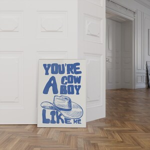 Puede incluir: Una impresi&oacute;n art&iacute;stica enmarcada con el texto "YOU'RE A COWBOY LIKE ME" en azul, con una ilustraci&oacute;n de un sombrero de vaquero. La impresi&oacute;n se muestra contra una pared blanca y un suelo de madera.