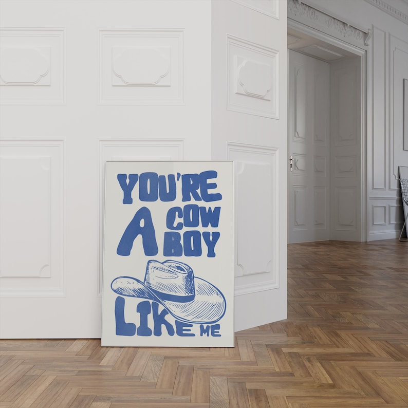 Puede incluir: Una impresi&oacute;n art&iacute;stica enmarcada con el texto "YOU'RE A COWBOY LIKE ME" en azul, con una ilustraci&oacute;n de un sombrero de vaquero. La impresi&oacute;n se muestra contra una pared blanca y un suelo de madera.
