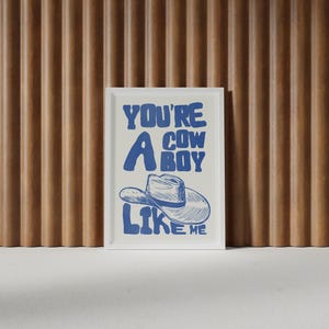 Puede incluir: Un estampado enmarcado con el texto "YOU'RE A COWBOY LIKE ME" en azul. La obra de arte presenta una ilustraci&oacute;n de un sombrero de vaquero. El estampado se muestra contra una pared de listones de madera.