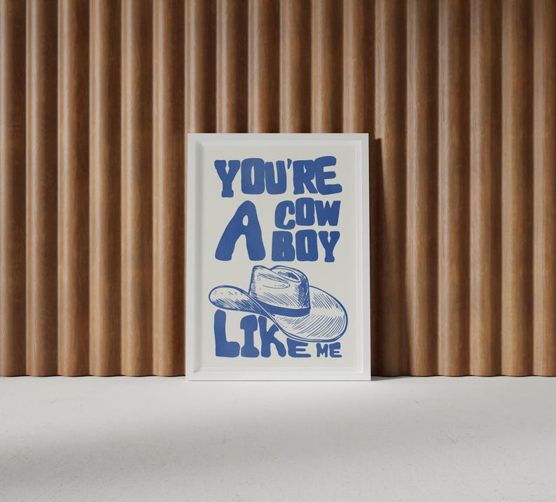 Puede incluir: Un estampado enmarcado con el texto "YOU'RE A COWBOY LIKE ME" en azul. La obra de arte presenta una ilustraci&oacute;n de un sombrero de vaquero. El estampado se muestra contra una pared de listones de madera.