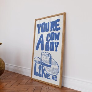 Puede incluir: Un cuadro enmarcado con el texto "YOU'RE A COWBOY LIKE ME" en azul, con una ilustraci&oacute;n de un sombrero de vaquero. La obra tiene un fondo blanco y un marco de madera, apoyado contra una pared blanca sobre un suelo de madera.