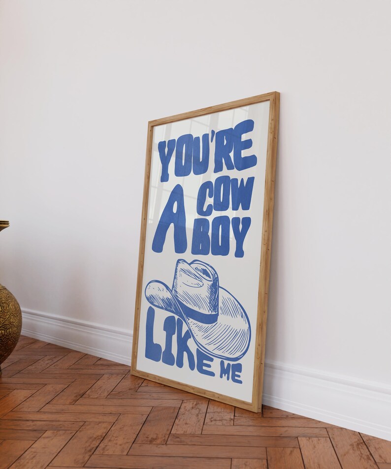 Puede incluir: Un cuadro enmarcado con el texto "YOU'RE A COWBOY LIKE ME" en azul, con una ilustraci&oacute;n de un sombrero de vaquero. La obra tiene un fondo blanco y un marco de madera, apoyado contra una pared blanca sobre un suelo de madera.