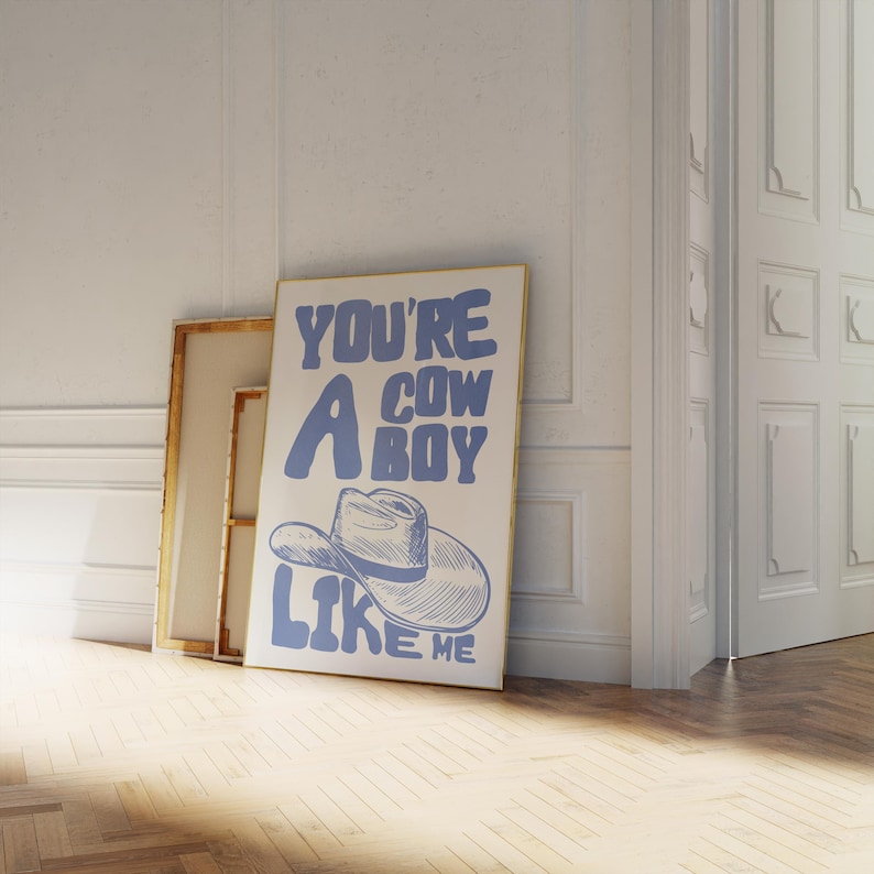Puede incluir: Un p&oacute;ster enmarcado con el texto "YOU'RE A COWBOY LIKE ME" en azul, con una ilustraci&oacute;n de un sombrero de vaquero. El p&oacute;ster se apoya en una pared blanca con otras obras de arte enmarcadas, sobre un suelo de madera.