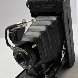 Può includere: Una macchina fotografica pieghevole vintage nera con una tracolla in pelle e un obiettivo contrassegnato "Ilex Anastigmat".