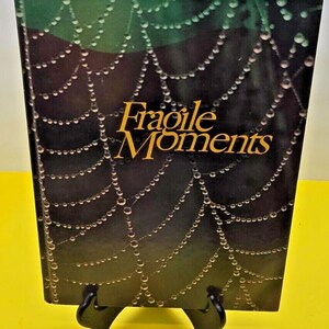 Könnte beinhalten: Ein Buch mit dem Titel "Fragile Moments" mit schwarzem Hintergrund und einem Design aus einem Spinnennetz mit Tautropfen.
