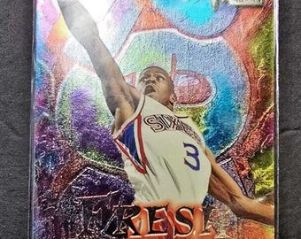 May include: A silver foil trading card featuring a basketball player in a white jersey with blue and red accents. The player is jumping in the air with the ball in his hand. The card has a holographic background with a rainbow effect. The text "Fresh Revelation" and "Allen Iverson" is printed on the card.