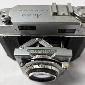 May include: A silver and black Ansco K-Promat 35mm film camera with a lens that has a depth of field scale and a focus ring. The camera has a black leather covering and a silver metal body.